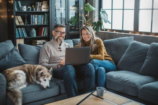 A Jerry Kelly Heating and Air Conditioning customer couple sits on a gray couch with their dog, reviewing services on a laptop at home.