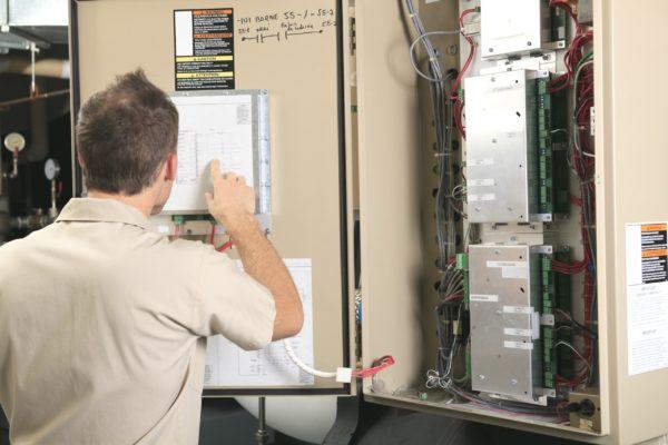A Jerry Kelly Heating and Air Conditioning technician reviews a schematic inside an open electrical panel with exposed wiring.