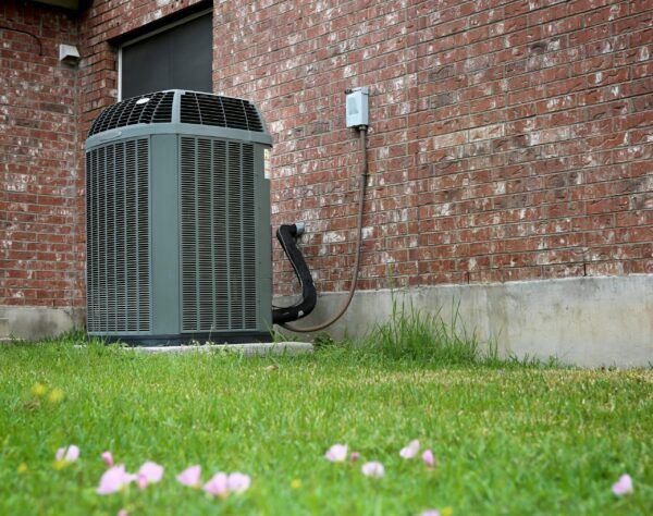 A Jerry Kelly Heating and Air Conditioning unit sits by a brick wall, surrounded by green grass and cheerful pink flowers in front.