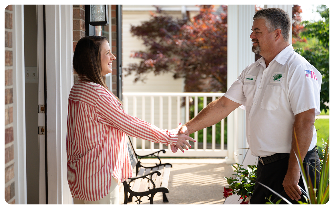 A woman and a man wearing a uniform shake hands on a porch outside a house on a sunny day.
