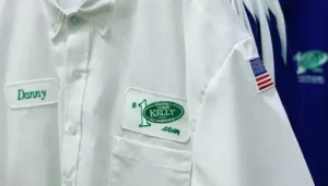 A white work shirt with green embroidered patches reading "Danny" and "Hey Kelly Heating & Air Conditioning", and a small USA flag patch on the sleeve.