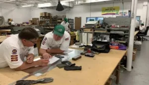 Two men in work uniforms discuss paperwork at a large workshop table; various tools and supplies are visible in the industrial workspace.