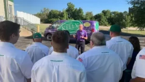 A man stands and speaks to a group of people in white shirts outdoors, with a campaign van displaying "Kelly" parked in the background.