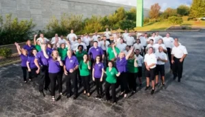 A large group of people poses outdoors in matching purple, green, and white shirts, standing in a parking lot with greenery and a concrete wall in the background.