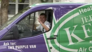 A man in a white shirt waves from the driver’s seat of a purple and green van labeled “Ask for: THE PURPLE VAN.”.