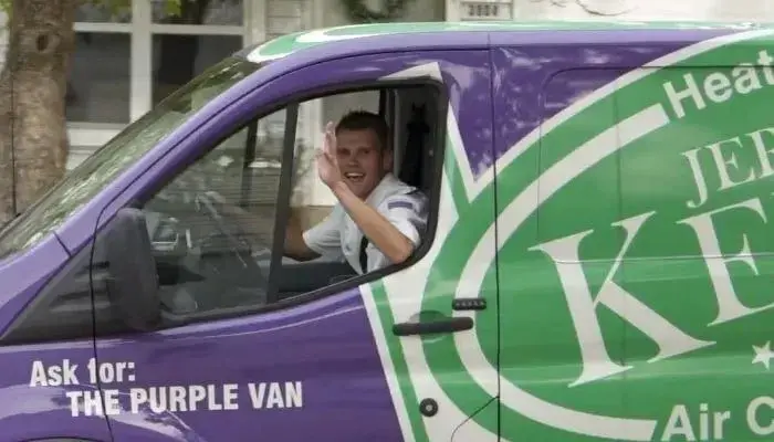 A man in a white shirt waves from the driver’s seat of a purple and green van labeled “Ask for: THE PURPLE VAN.”.