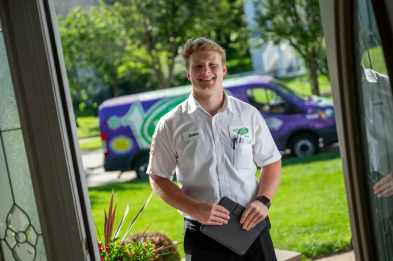 A Jerry Kelly technician in a crisp white uniform stands at a customer's door holding a tablet, with our branded van outside amid greenery.