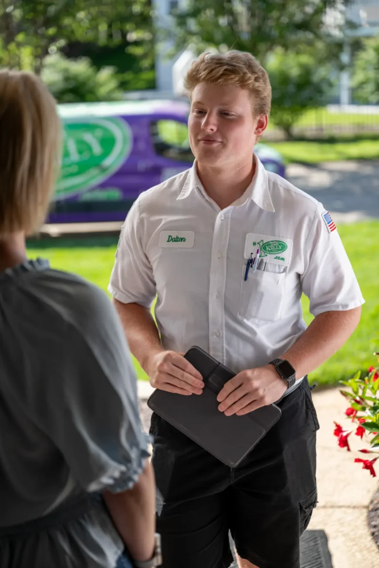 A Jerry Kelly technician chats with a customer at their door, tablet in hand, as the company van sits parked on a suburban street.