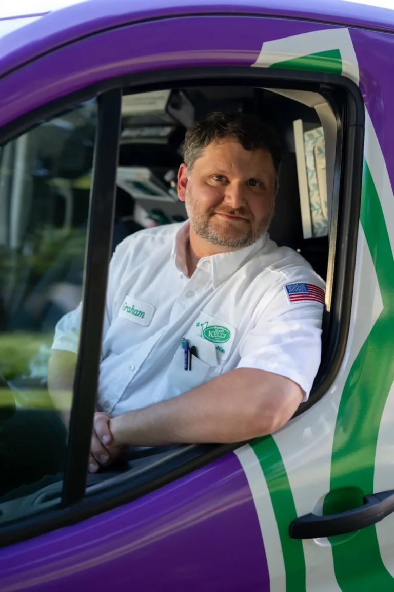 A man in a white uniform shirt with a name tag sits in the driver's seat of a purple and green vehicle, looking at the camera.